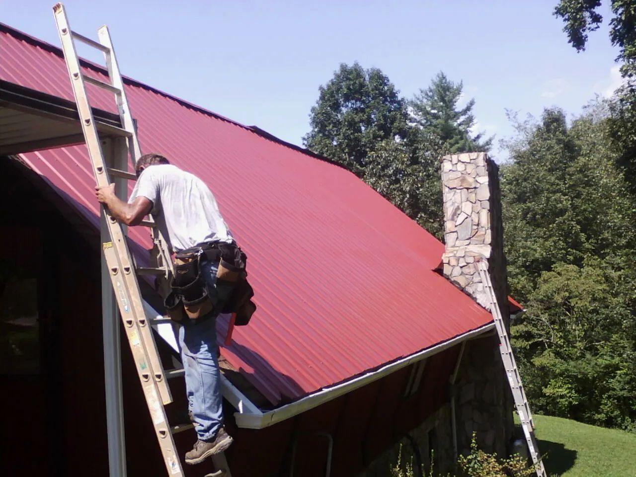 Roofer on a ladder beside a red metal roof and stone chimney — Soffit & Fascia Repair by Roofing Brothers in Fultondale
